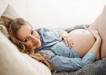 Młoda kobieta w pozycji półsiedzącej obejmuje dłońmi odłonięty ciążowy brzuch.