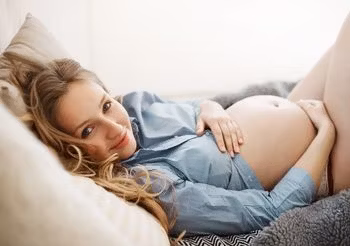 Młoda kobieta w pozycji półsiedzącej obejmuje dłońmi odłonięty ciążowy brzuch.